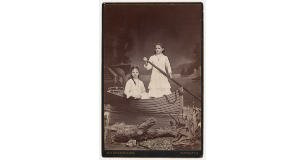 A sepia-toned studio photograph of two adolescent White girls, both wearing high-necked white dresses, posing in a rowboat in front of a painted lake scene.
