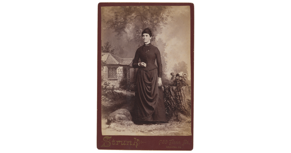A sepia-toned studio photograph of a White woman with dark hair wearing a corseted, high-necked dress standing in front of a painted pastoral scene with a cottage.