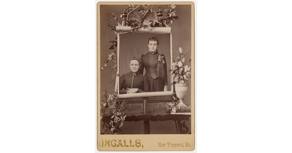 A sepia-toned studio portrait photograph of two White women on a massive paper scroll mounted on an easel and surrounded by flowers.