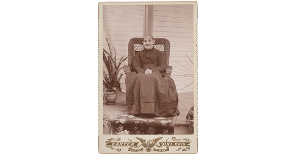 A sepia-toned studio photograph of an elderly White woman wearing a dark, high-necked dress sitting in a high back wicker chair on a porch with plants.