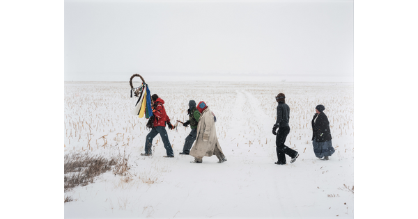 A color photograph of a group of people crossing a vast, snowy landscape.
