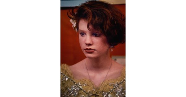 A color portrait photograph of a young White girl with short red hair, a flower tucked over one ear, and a decorative yellow top.