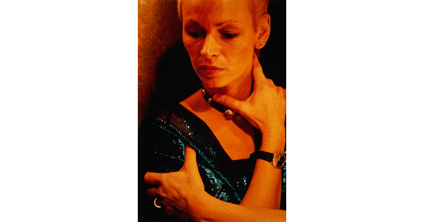 A color portrait photograph of a White woman, from forehead to upper arms, wearing a shimmering blue top, one arm wrapped around an upper arm, the other reaching up to her neck.