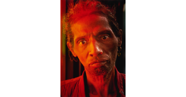 A color portrait photograph of a Black man cast in red light, hair combed back and stubble on his face, hoop earrings in one ear.