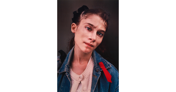A color portrait photograph of a young fair-skinned woman with brown hair, wearing a light pink cardigan under jean jacket with a red ribbon pinned to the collar.