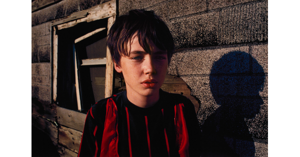A color portrait photograph of a White boy with brown hair wearing a red and black striped shirt, standing in front of a building partially covered in roof shingles.