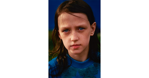 A color portrait photograph of a young White child with freckles, shoulder length brown hair, and blue eyes, wearing a blue knit sweater.