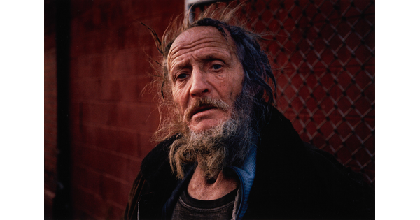 A color portrait photograph of an older White man with flyaway and matted hair, a graying beard and mustache, and a furrowed brow standing in front of a chain link fence.