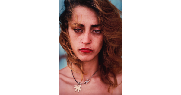 A color portrait photograph of a White woman with brown eyes, wavy shoulder-length brown hair, a gold stud in one nostril, red lips, and wearing a hemp leaf necklace.