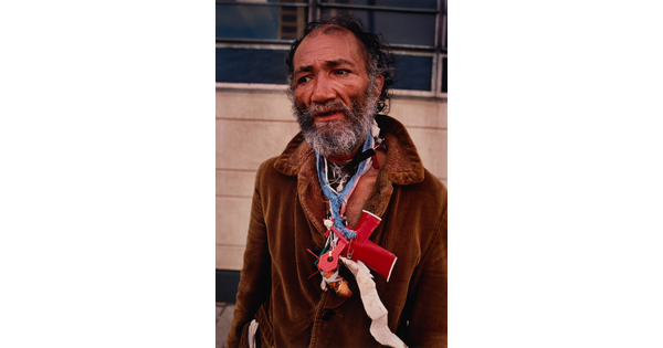 A color portrait photograph of a balding Black man with graying beard wearing a brown coat and random objects around his neck.