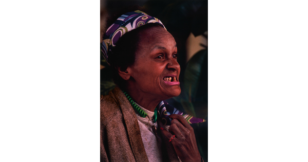 A color portrait photograph of a Black woman, smiling so that her underbite and missing teeth show, wearing a colorful scarf in her hair.