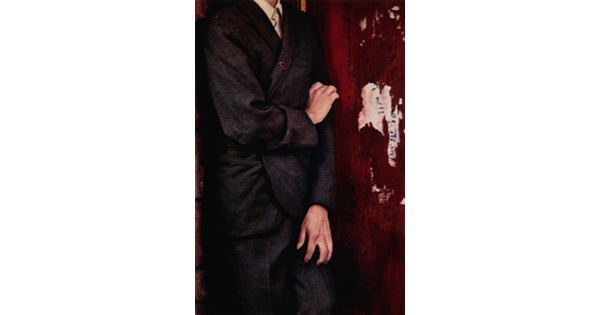 A color photograph of a White person from the neck down, wearing a loose-fitting suit, clawing at their leg and arm with their hands.