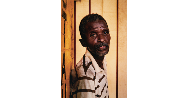A color portrait photograph of an older Black man, short hair and stubble on his thin face, wearing a striped polo shirt, and leaning against a wall.