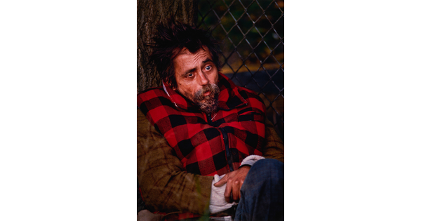 A color portrait photograph of a White man, seated against a tree, with messy dark hair, misshapen eyes and a graying goatee, wearing a red-and-black checked vest.