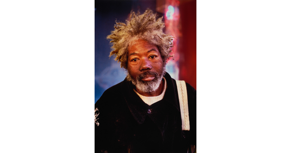A color portrait photograph of an older Black man with matted gray hair and a beard wearing a black jacket.