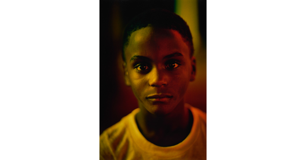 A color portrait photograph portrait of a young Black person, close-cropped hair, wearing a white t-shirt; yellow lighting casts tinted shadows across their face.