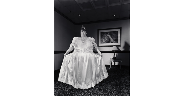 A black-and-white photograph of a White woman holding out the skirt of her long, formal white dress.