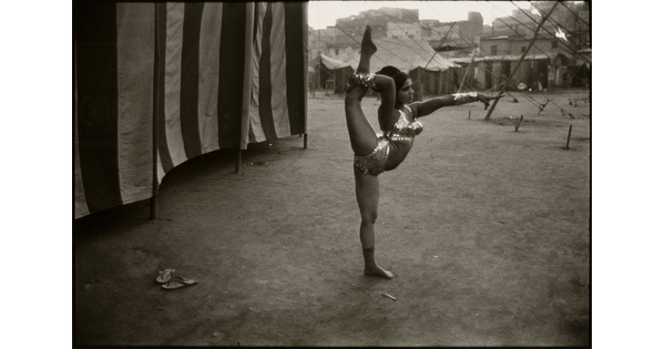 A black-and-white photograph of a dark-skinned woman with dark hair in a sequined bikini and metallic cuffs stretching her leg behind her head as she stands next to a striped tent.