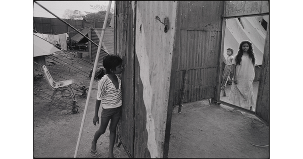 A black-and-white photograph of a dark-skinned boy peeking through a hole in a wooden structure as two dark-skinned girls appear in a doorway on the opposite side.