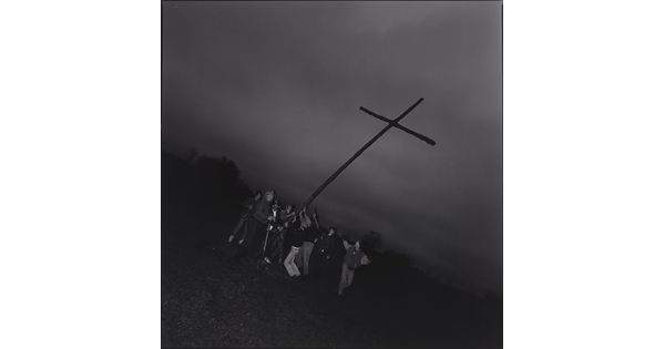 A black-and-white photograph of a group of people hoisting up a large cross in a field at dusk.