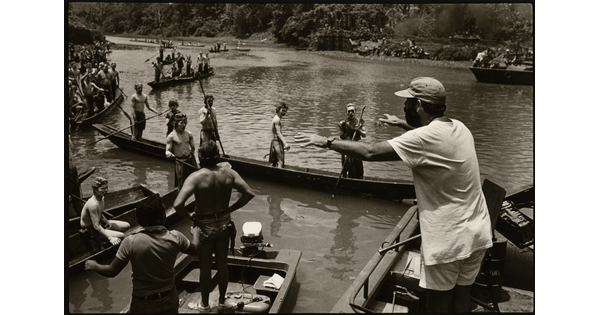 A black-and-white photograph of a man addressing a group of people in boats on a river.