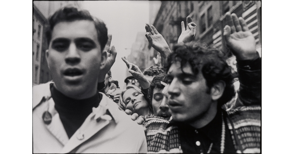 A black-and-white photograph of two people amongst a crowd of protesters, some of whom are holding up their hands and making circles with their index fingers and thumbs.