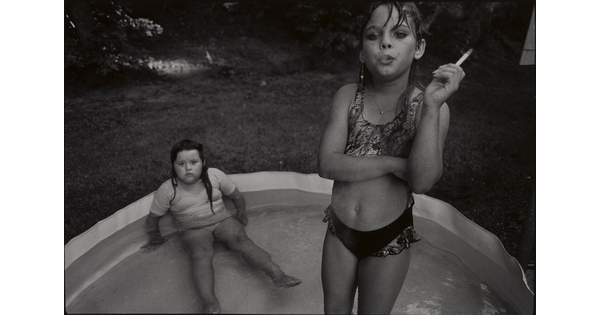 A black-and-white photograph of a young White girl standing in a kiddie pool smoking a cigarette as another young White girl sits in the pool behind her.