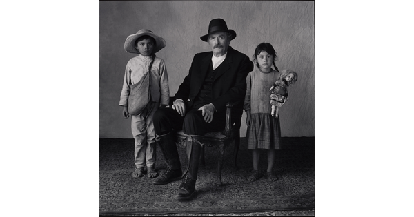 A black-and-white portrait photograph of a seated White man with a moustache, wearing a hat, black suit, and tall boots with two medium-skinned children standing on either side of him.