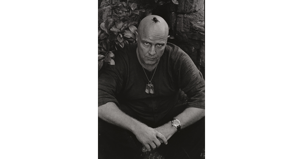 A black-and-white photograph of a bald White man seated on the ground with his arms resting on his knees and a large beetle on his head.