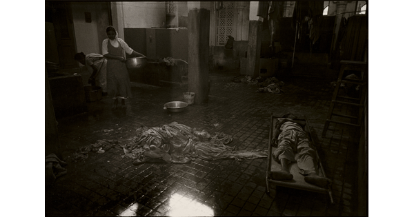 A black-and-white photograph of the inside of a room showing a pile of rags on a wet tile floor, people on one side of the room holding tubs, and a person lying on a cot on the other side.