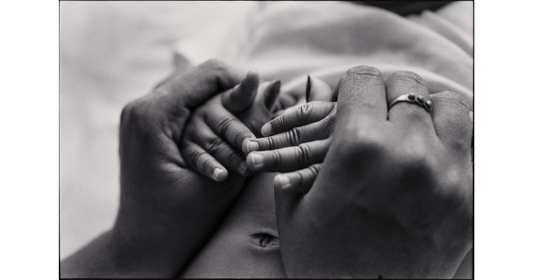 A close-up, black-and-white photograph of adult Black hands clasping Black baby hands.