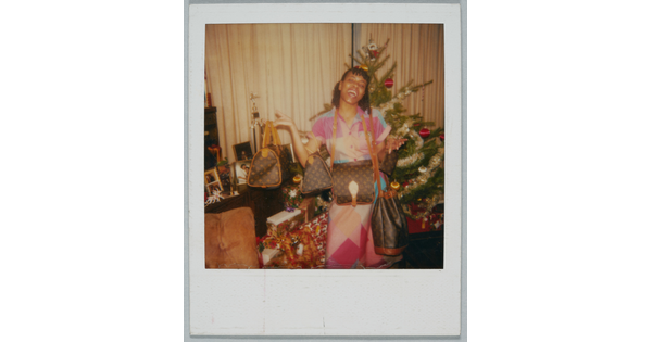A color Polaroid photograph of a Black woman gleefully showing off several designer purses in front of a Christmas tree.