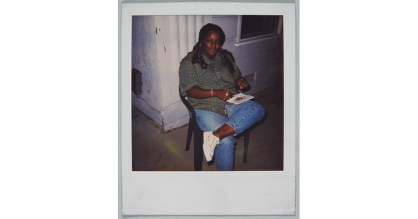 A color Polaroid photograph of a Black woman seated on a plastic chair balancing a piece of paper with bits of something on it on her crossed leg.