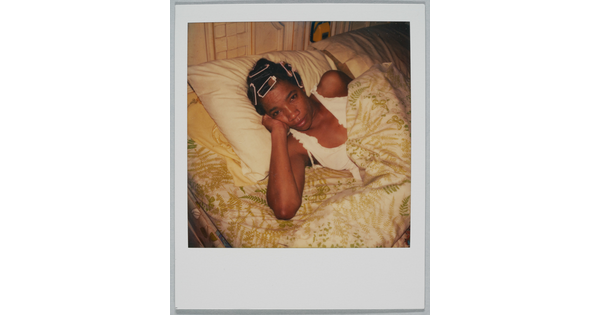 A color Polaroid photograph of Black woman, curlers in her hair, in bed with her head propped up on her hand.