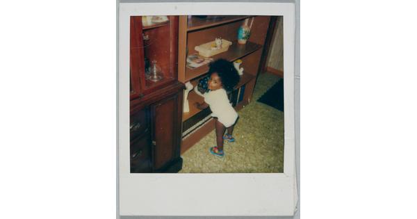 A color Polaroid photograph of Black toddler in a shirt, diaper, and sandals with big, natural hair propping themselves up on a bookshelf.