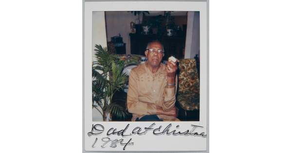 A color Polaroid photograph of a seated, middle-aged Black man in glasses and a light brown shirt holding an apple in one hand; handwritten text at bottom reads: "Dad at Christma[s] 1984."