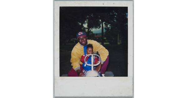 A color Polaroid photograph of a Black man and toddler seated behind the handles of an outdoor play toy in a park.