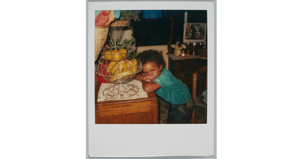 A color Polaroid photograph of a Black child leaning on a table that has a tiered fruit holder.
