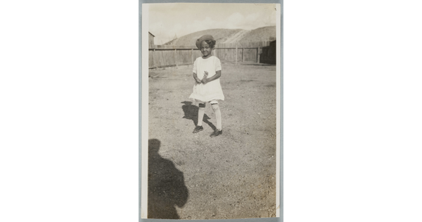 A black-and-white snapshot of a Black girl in a white dress and knee socks standing in a fenced yard.