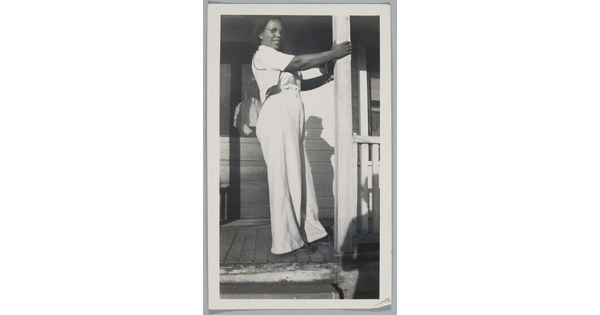 A black-and-white snapshot of a Black woman in white shirt and pants standing on a porch holding onto a column.