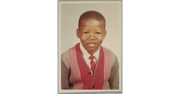 A color school photograph of a young, smiling Black boy wearing a white collared shirt and red tie under a striped cardigan.