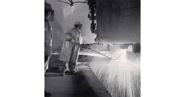 A black-and-white photograph of an industrial worker, wearing safety helmet, coat, and gloves holding a rod under a machine emitting sparks.