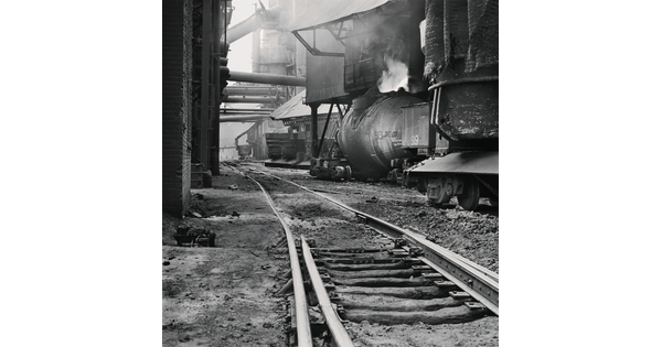 A black-and-white photograph of railroad track  between industrial buildings and  machinery, some emitting steam or smoke.
