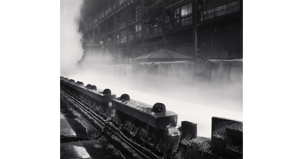 A black-and-white photograph of industrial machinery in front of steam or smoke rising near a multi-story industrial building.