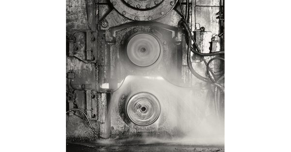 A black-and-white photograph of two spinning metal discs set into a metal wall that has pipes and tubes connected to it.