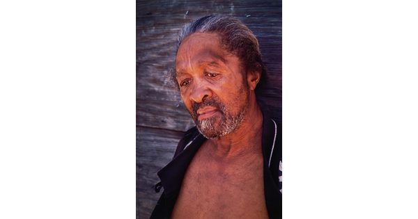 A color portrait photograph of a Black man, salt-and-pepper hair slicked back, gray beard, downward gaze, wearing a black jacket open to reveal his bare chest.