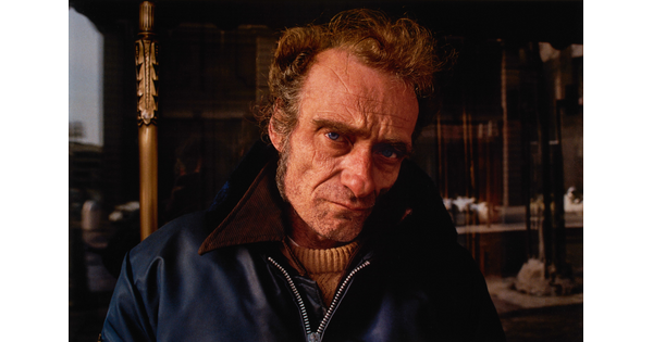 A color portrait photograph of a White man with a wrinkled face, thick brows, long sideburns, and sunken cheeks wearing a blue jacket with a tan collar.