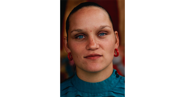 A color portrait photograph of a White person with light blue eyes, hair pulled back tightly on their head, wearing red hoop earrings, and a teal top.