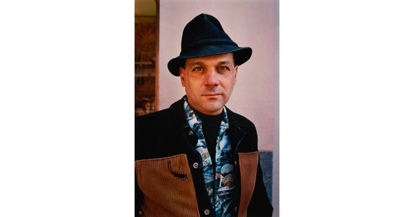 A color portrait photograph of a fair-skinned man wearing a fedora hat and a blue Hawaiian print shirt under and black and brown jacket.