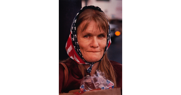 A color portrait photograph of a middle-aged White woman with shoulder-length hair, prominent cheekbones and creases around her eyes, wearing an American flag scarf around her head.